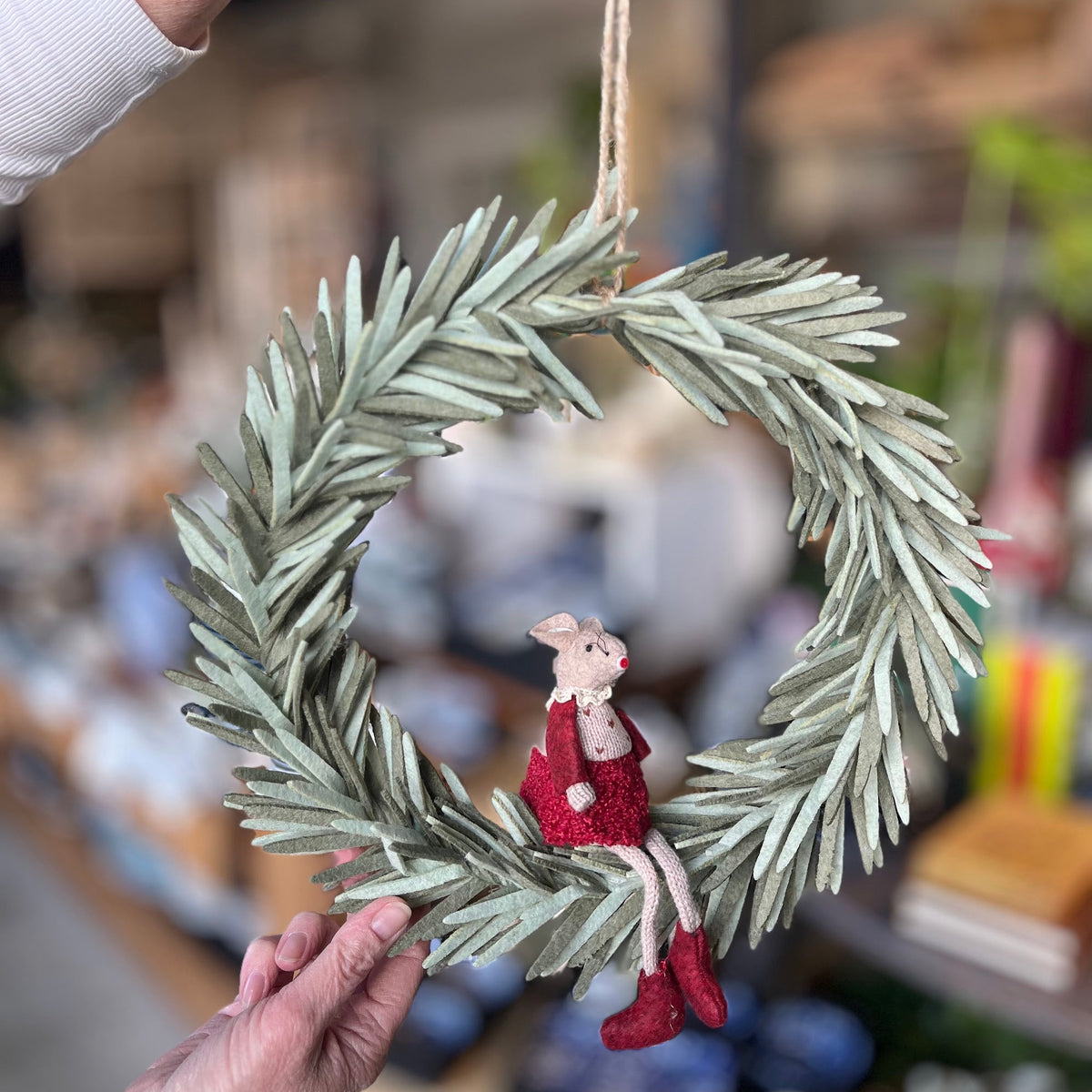 A rustic olive-green pine wreath ornament with a small seated rabbit figurine in a red dress and red boots, held by hands in a blurred shop setting.