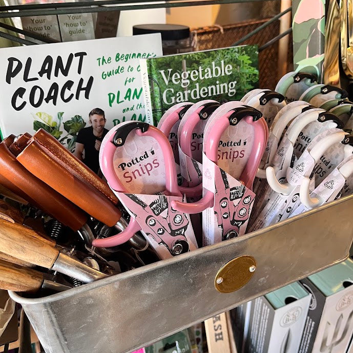A metal storage bin filled with gardening tools, including pink and white hand prongs labeled 'Potted's Snips,' alongside gardening books such as 'Plant Coach' and 'Vegetable Gardening.'.