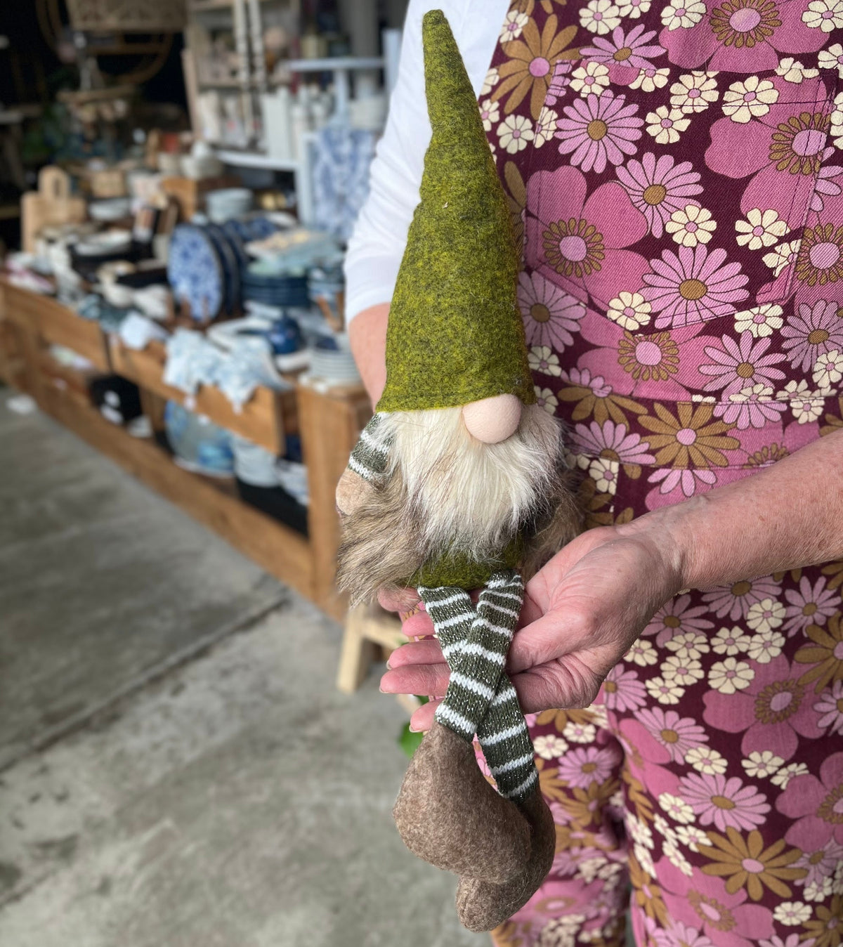 A person in a pink floral apron holds a small gnome doll with a green felt hat, white beard, and striped green-and-white scarf in a rustic shop setting.