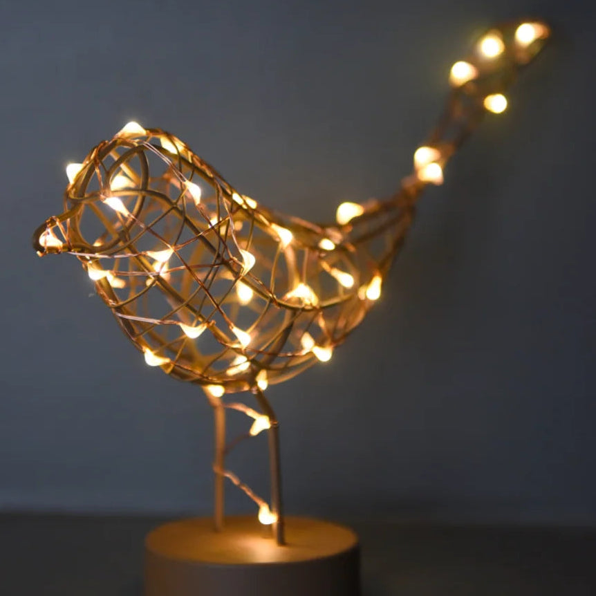 A decorative bird-shaped wire sculpture illuminated with warm white fairy lights, mounted on a round wooden base against a dark gray background.
