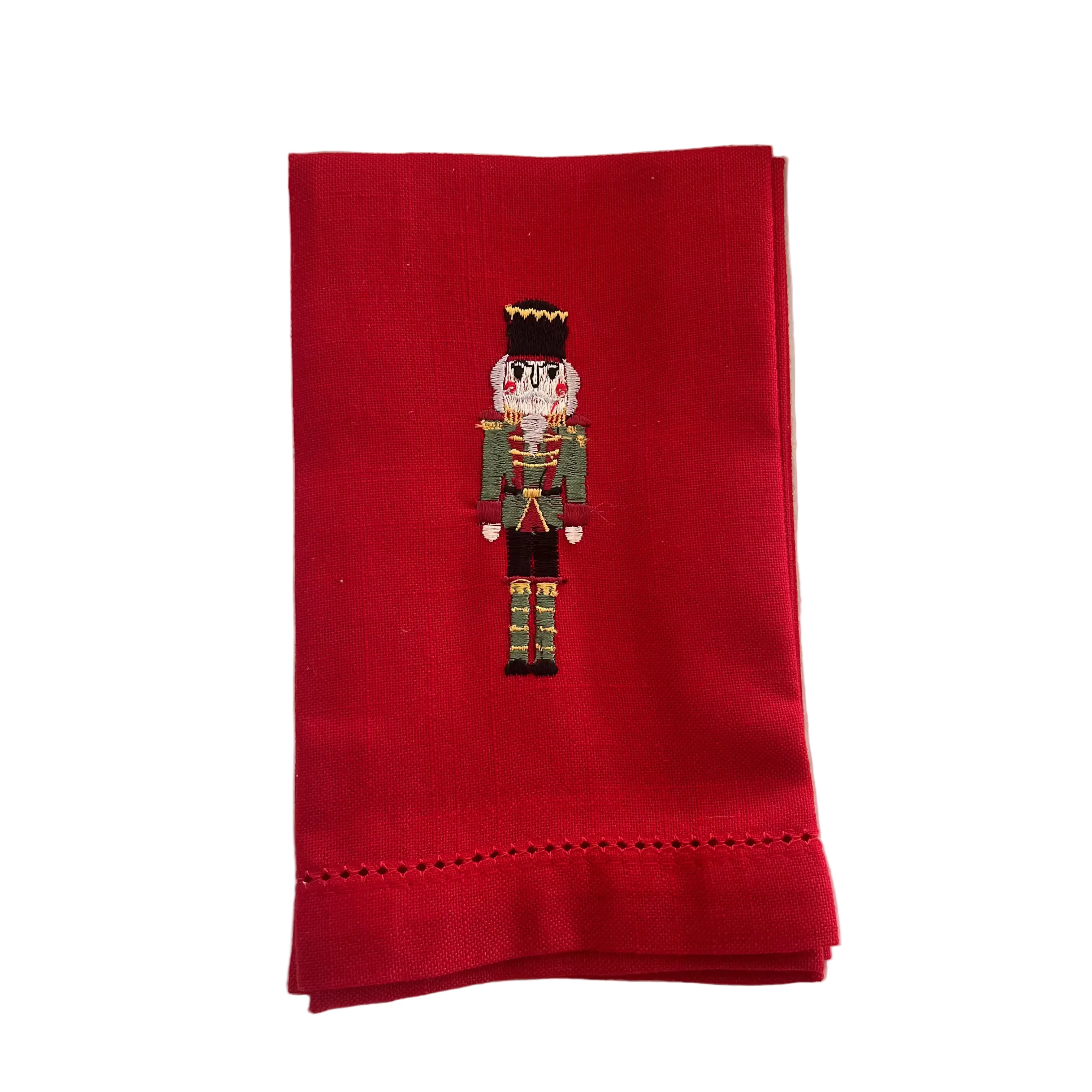 A folded red fabric napkin with a small embroidered nutcracker figure in the center and decorative hemstitching along the bottom edge, shown on a plain white background.