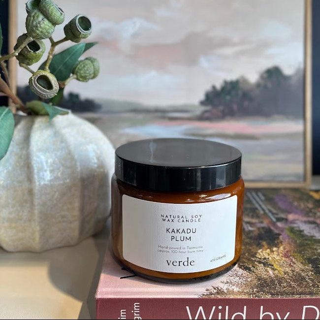 A jar candle labeled 'KAKADU PLUM' by Verde sits on a book, styled with a white textured vase containing eucalyptus branches and a framed landscape painting in the background.