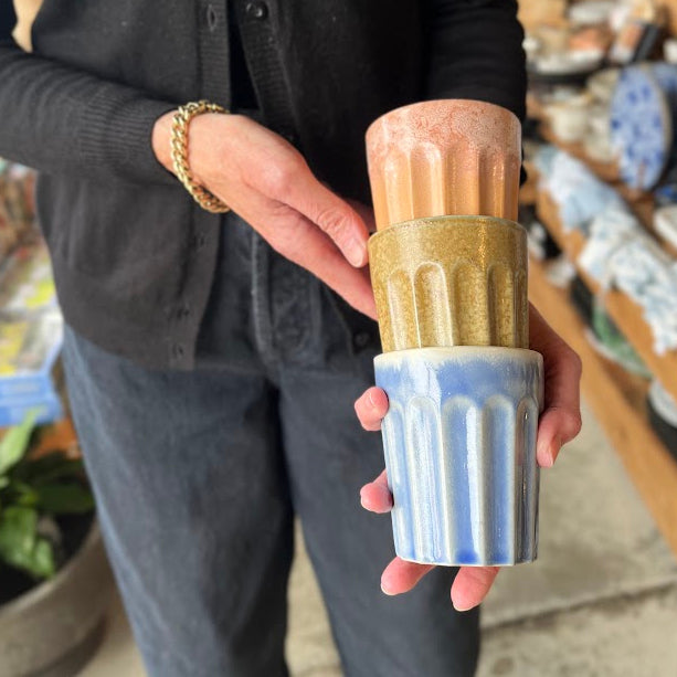 A person wearing a black cardigan and gray pants holds a stack of three ribbed ceramic cups with vertical fluting. The cups have different glazes: a terracotta orange top, a textured gold middle, and a light blue bottom.