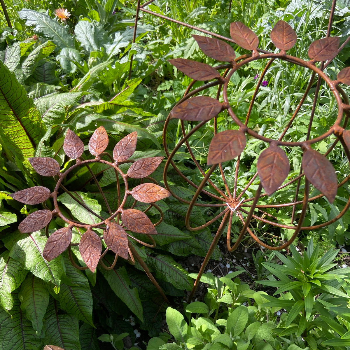 A rust-colored metal poppy garden stake, shown planted in a lush green garden.
