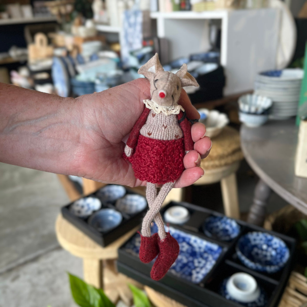 A handmade knitted mouse doll with a red dress and red shoes is held in a person's hand, with a pottery and tableware shop blurred in the background.
