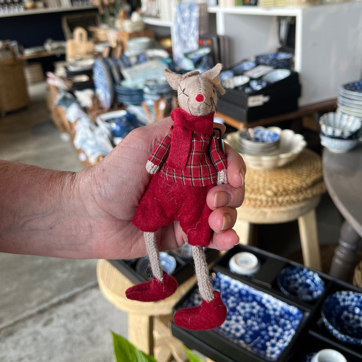 A hand holds a small mouse dressed in red plaid and red shoes inside a home goods or pottery shop with blue-and-white ceramics in the background.