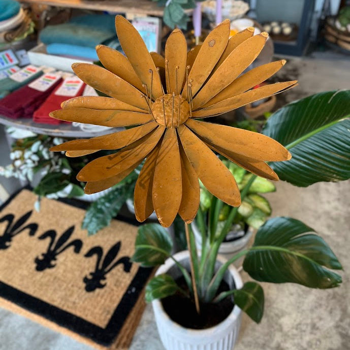 A large rusty flower decoration with mustard-yellow petals and green leaves, displayed in a white ribbed pot on a concrete floor in a shop setting.