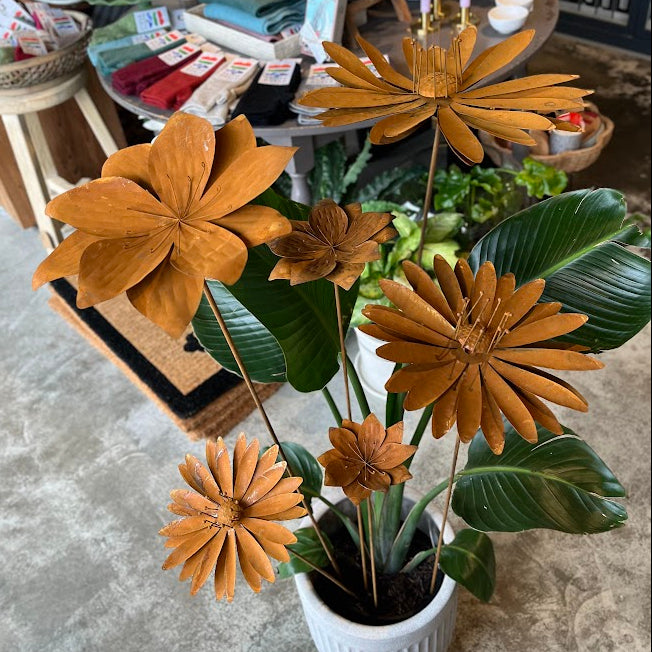 A potted decorative artificial plant with large rusty flower heads and glossy green leaves in a white ribbed pot, photographed indoors on a concrete floor with a shop display in the background.