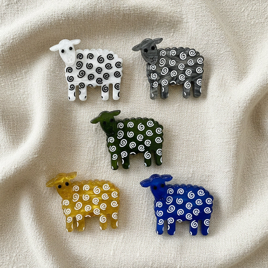 Five small sheep brooches in white, gray, olive green, yellow, and blue, each covered with white spiral patterns, arranged on a plain white background.