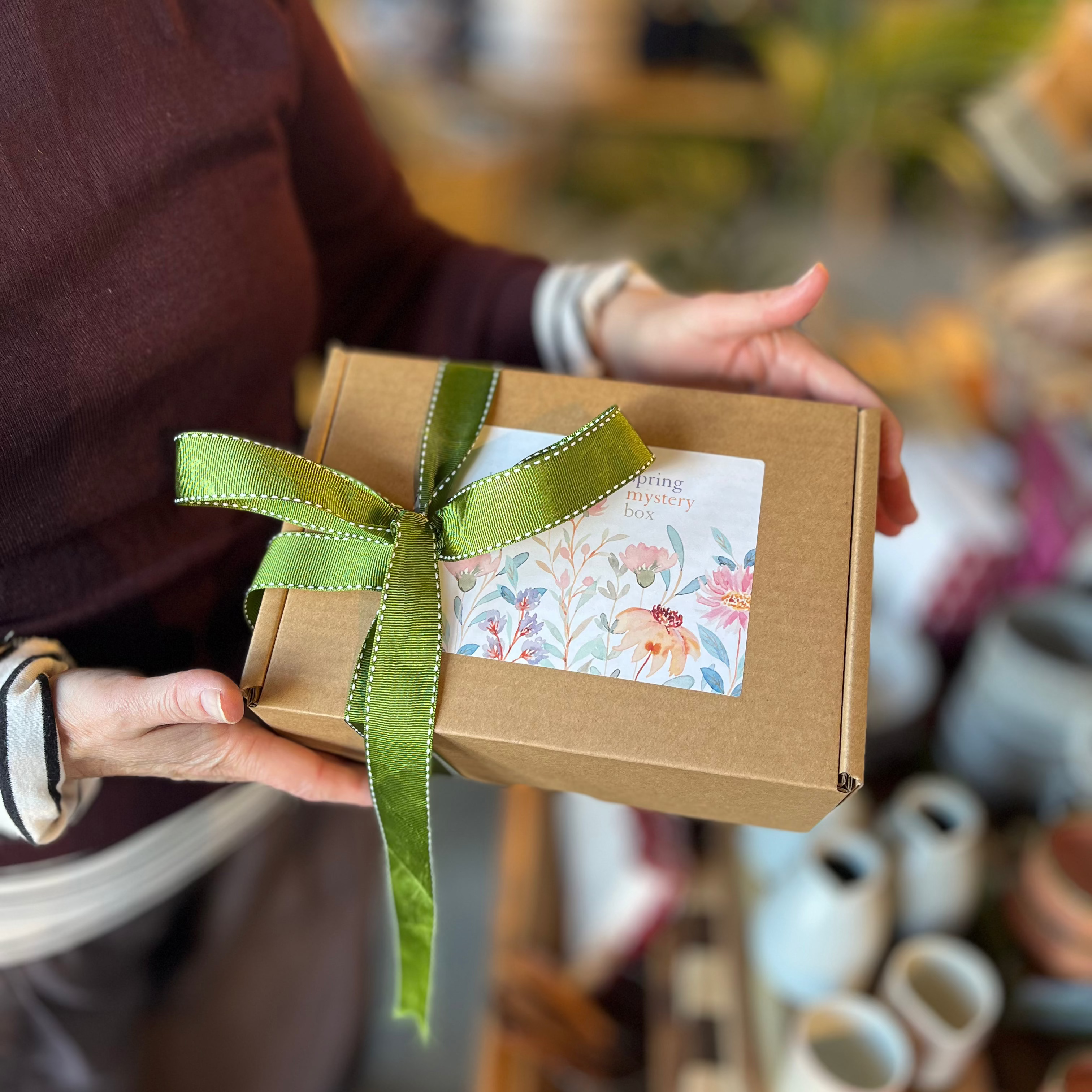 A person holding a small brown kraft gift box tied with a green ribbon and a floral label, photographed in a softly blurred shop or home setting.