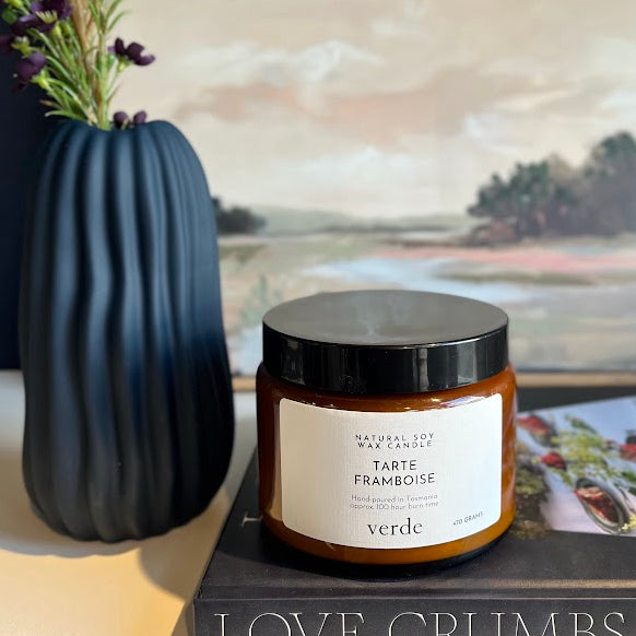 A jar candle labeled TARTE FRAMBOISE by Verde sits on a book on a tabletop, styled with a dark blue ribbed vase holding lavender sprigs and a framed landscape print in the background.