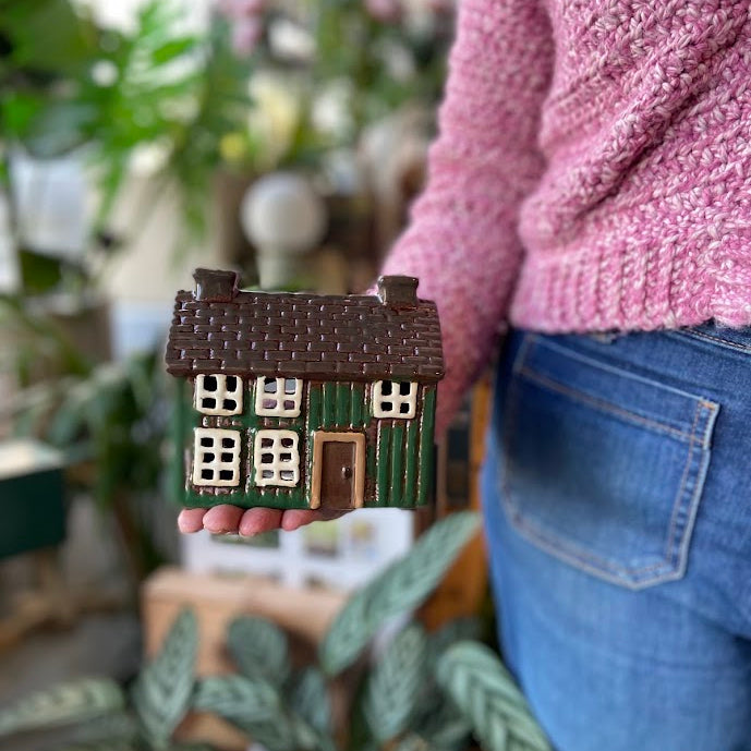A person in a pink knit sweater and blue jeans holds a small decorative house figurine with a brown roof, green walls, and cream window details against a softly blurred indoor background with plants.