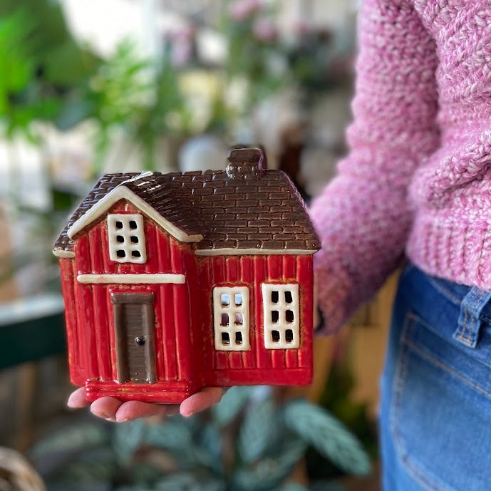 A person in a pink knit sweater and blue jeans holds a small red house-shaped ceramic ornament with a brown roof in a softly blurred indoor setting with plants.
