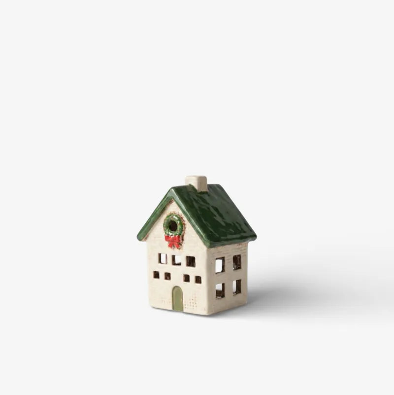 A small ceramic Christmas house ornament with a cream-colored body, a green roof, and a green wreath with a red bow on the front, set against a plain light gray background.