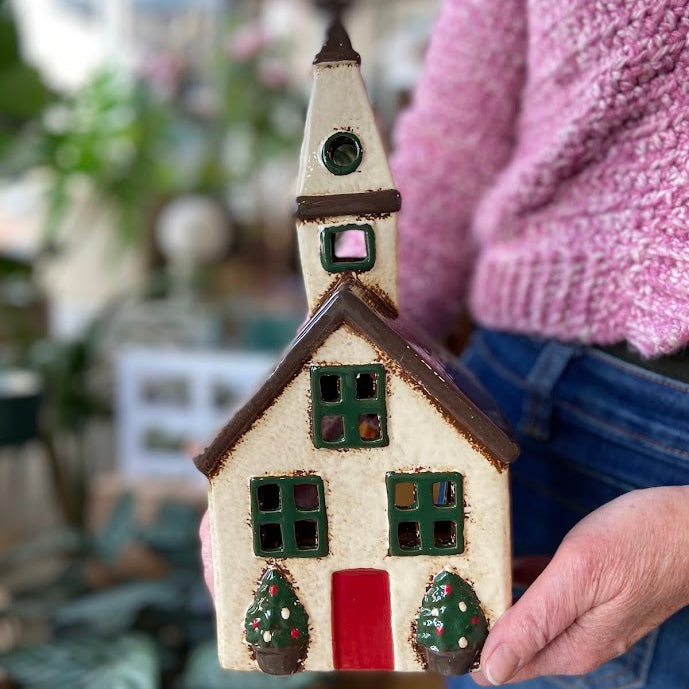 A hand holds a small decorative house figurine with a cream-colored body, brown roof, green window frames, a red door, and small green tree decorations, while a person in a pink sweater and blue jeans is partially visible in the background.