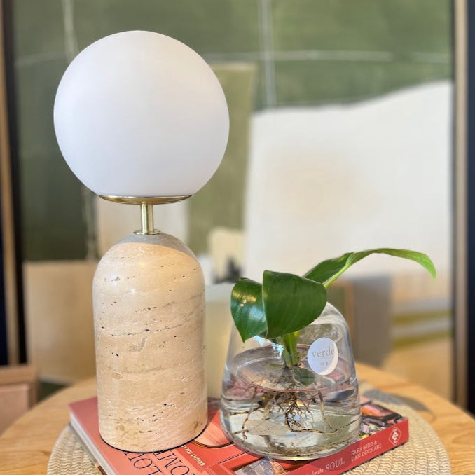 Decorative travertine lamp with a spherical white shade on a textured beige base, placed on a book with a plant in a glass vase.