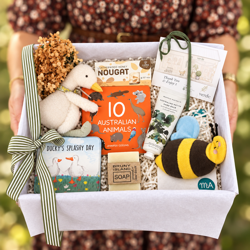 A baby hamper filled with baby and animal-themed items, held open by hands wearing a floral top. The baby hamper box contains a plush duck toy, a bumblebee plush, a soap bar, a card, and other baby accessories.