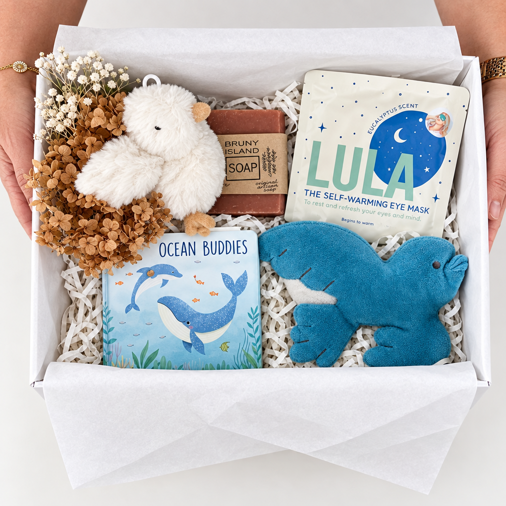 A curated gift box filled with bath and beauty items, including a blue whale card, a blue plush bath ornament, a white eye mask packet labeled LULA, a plush white bird, and dried orange flowers, held open by hands over a white surface.