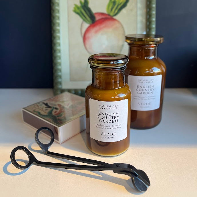 Two amber glass jars of English Country Garden  candles on a tabletop, with a black metal tongs tool in front, a decorative box, and a framed botanical print in the background.
