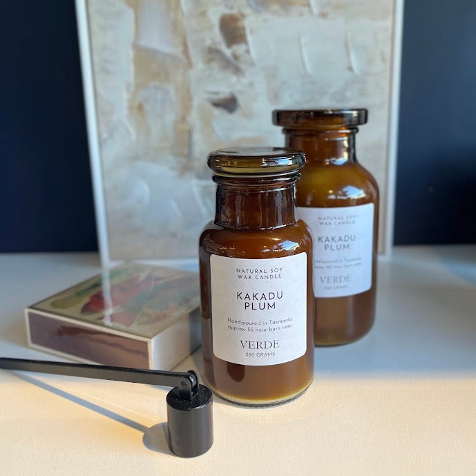 Two amber glass candle jars labeled 'KAKADU PLUM - VERDE' with white labels, shown on a tabletop with a matchbox and candle wick in front, and a framed abstract print against a dark blue background.