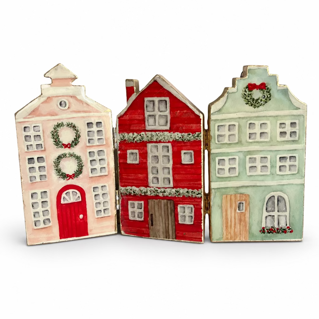 Three small hand-painted house-shaped ornaments or decorative panels standing side by side on a reflective white surface. The left house is pale pink with a red door and green wreath; the middle is red with white trim; the right is pale green with a wreath and brown door.