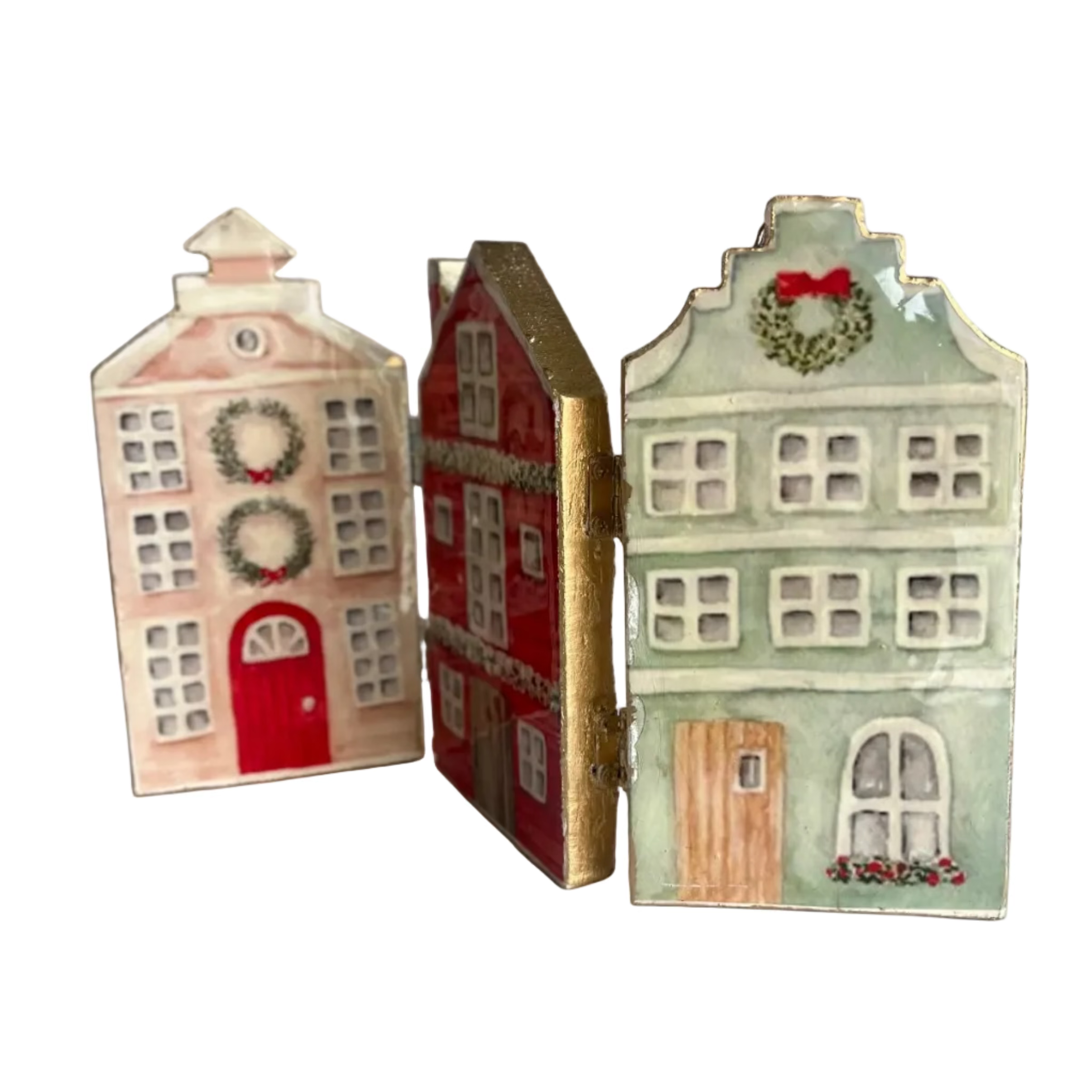 Three small decorative house-shaped ornaments or boxes with a vintage holiday design, standing upright on a white surface. One is light pink with a red door and wreath, one is red with gold trim, and one is pale green with a wreath and red bow.