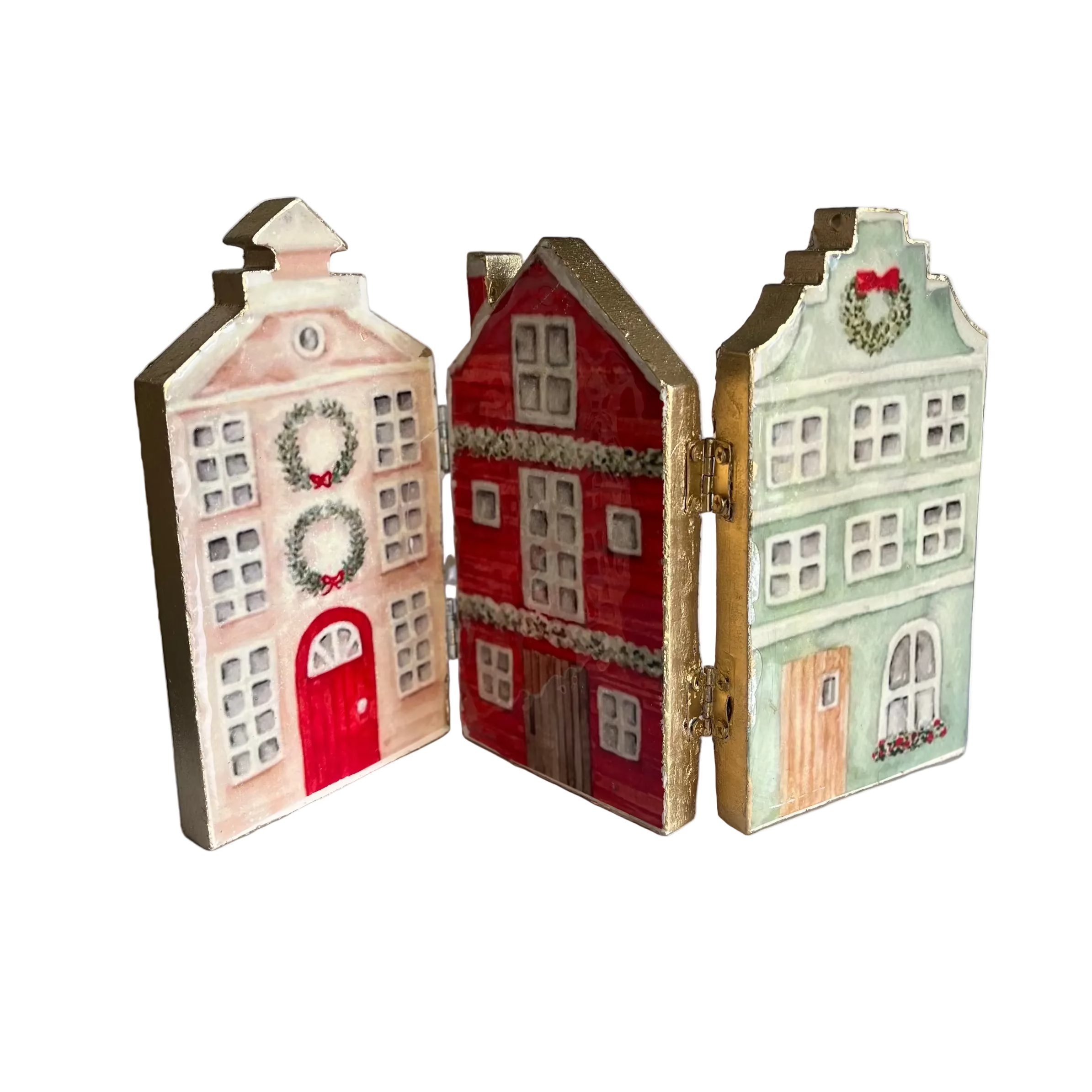 Three hand-painted wooden Christmas house ornaments displayed side-by-side on a white surface. The left ornament is light pink with a red door and wreath; the middle ornament is red with gold trim and white window details; the right ornament is light green with gold trim and a wreath on the gable.