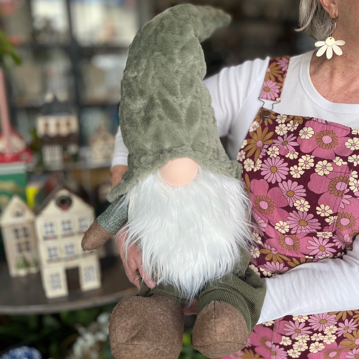 A person in a pink floral apron holds a plush gnome doll with a green hat, white beard, and brown shoes in a shop setting.