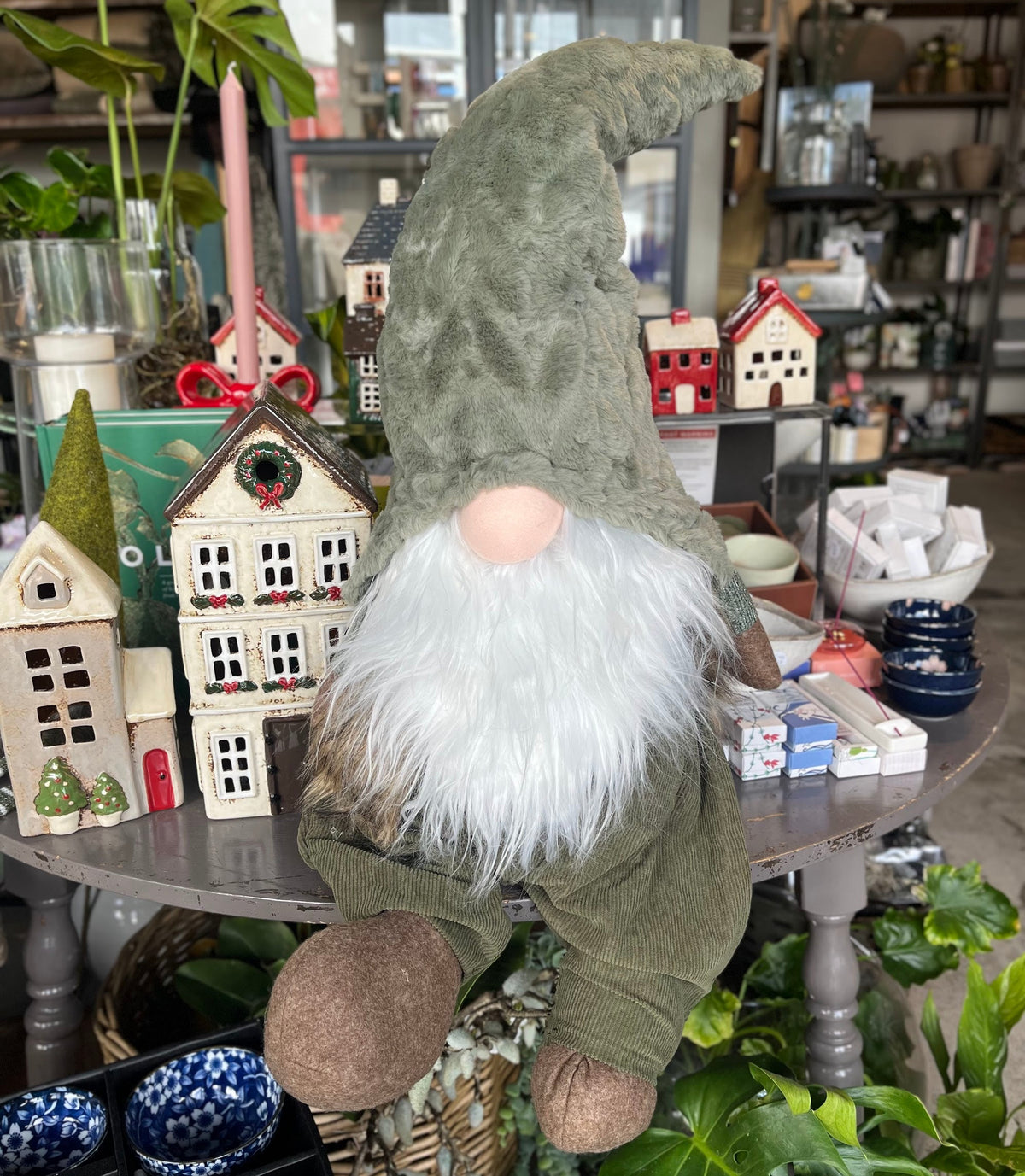 A plush gnome decoration with an olive green hat and body, white beard, and brown shoes, displayed on a table surrounded by miniature houses and plants in a shop setting.