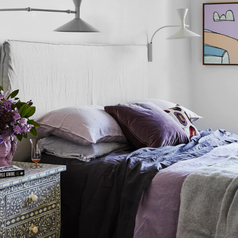 linen sheets in purple, lilac, navy styled on a bed in natural light with a white bedhead